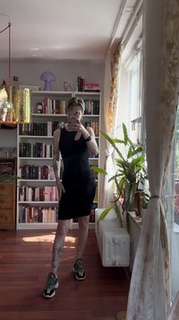 Černé bezrukávové šaty s výřezem; přiléhavý střih. Nosené s vícebarevnými teniskami. V pozadí knihovny a rostliny.