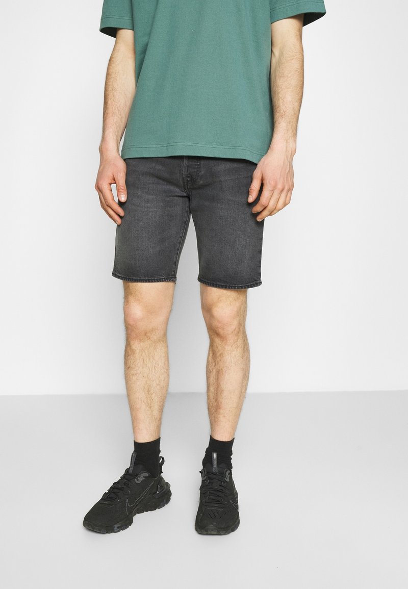 Homme portant un short en denim noir délavé, un t-shirt vert, des chaussettes noires et des chaussures de sport noires, debout devant un fond blanc.