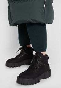 Botas de ante negras hasta el tobillo con una suela de plataforma gruesa, con cordones redondos y ojales de metal, combinadas con leggings verde oscuro y un abrigo acolchado.