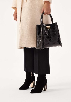 Borsa nera in pelle stampata coccodrillo, di forma quadrata, con finitura lucida e accessori in oro. Abbinata a pantaloni neri aderenti e stivaletti con tacco.