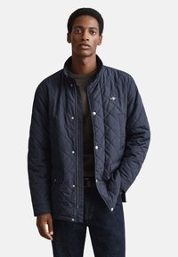 Námořnicky modrá prošívaná bunda s vysokým límcem, zapínáním na druky a logem na hrudi. Má boční kapsy a je nošena přes tmavý svetr.