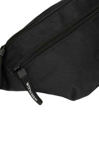Bandolera negra de tela con superficie texturizada, que cuenta con un cierre con cremallera y una etiqueta de marca, diseñada para un almacenamiento compacto y conveniencia.