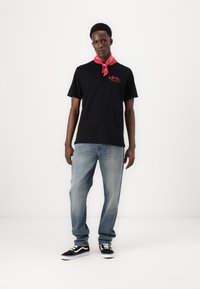 Zwart t-shirt met een rood logo, losse lichtblauwe jeans, zwart-witte sneakers en een rode bandana om de nek geknoopt.