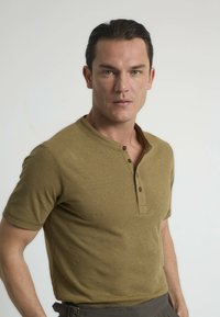 Camisa henley de manga corta, de color verde oliva, hecha de una tela suave, con una tapeta de tres botones y cuello acanalado.