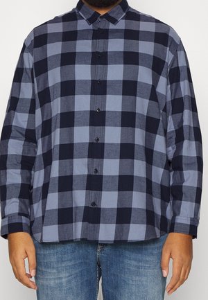 Homme portant une chemise à manches longues à carreaux bleus et noirs avec un jean en denim, debout devant un fond uni.