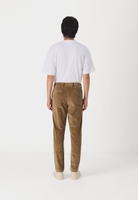 Pantaloni in velluto a coste marroni con una vestibilità slim, caratterizzati da tasche posteriori e una texture liscia, abbinati a una t-shirt bianca e scarpe chiare.