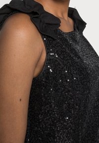 Svart paljettklänning med en texturerad tyg, volangdetaljer vid axlarna och rundad halsringning som framhäver glittrande detaljer.