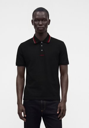 Bărbat purtând un tricou polo negru cu mâneci scurte, cu finisaj roșu la guler și un mic logo roșu „Versace” pe piept, stând în fața unui fundal alb.