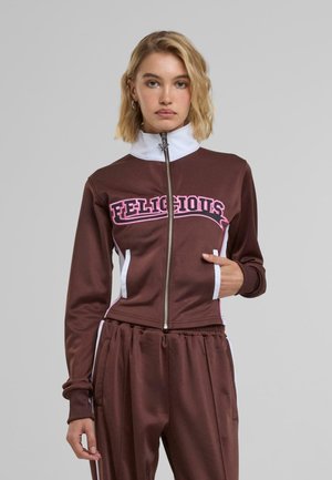 Bruine zip-up jas met witte accenten en "FELICIOUS" in roze letters. Glad textuur, hoge kraag en twee zijzakken aanwezig.