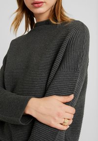 Dunkelgrauer rippierter Pullover mit hohem Kragen und langen Ärmeln. Der Stoff fühlt sich weich an und weist ausgeprägte vertikale Streifen auf.