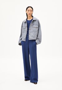 Chaqueta de mezclilla en azul claro, de corte oversize con dos bolsillos frontales, combinada con pantalones anchos de color azul oscuro y zapatos negros. Textura de tela suave.
