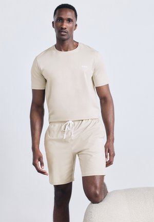 Muž stojaci s jedným kolenom opretým o štruktúrovanú bielu stoličku, oblečený v béžovej krátkej košeli a zodpovedajúcich krátkych nohaviciach so sťahovacou šnúrkou.