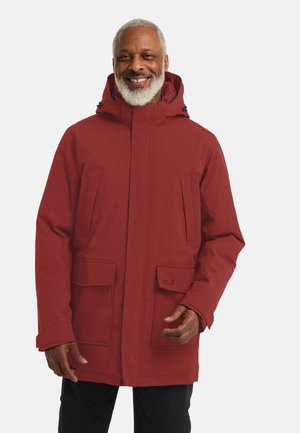 Lachende man met grijs haar en baard, gekleed in een rode winterjas met capuchon en zwarte broek, staand tegen een effen achtergrond.