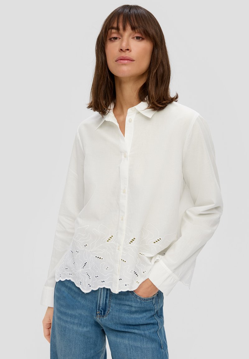 Weißes Hemd mit Knöpfen aus leichtem Stoff, verziert mit bestickten Blumenmustern und einer gewellten Saumkante, kombiniert mit Jeans.