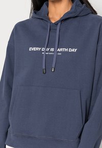 Mörkblå hoodie i bomull, med en framficka, dragsko i huvan, och vit text "VARJE DAG ÄR JORDENS DAG" på bröstet.