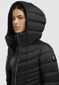 Sort puffer jakke med høj krave og stor hætte. Har lynlås lukning, struktureret quiltet mønster og et logo patch på ærmet.