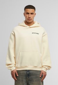 Haut à capuche couleur crème avec une poche avant, capuche réglable à cordon, et un logo "anomalie" vert sur la poitrine. Tissu doux, coupe décontractée.