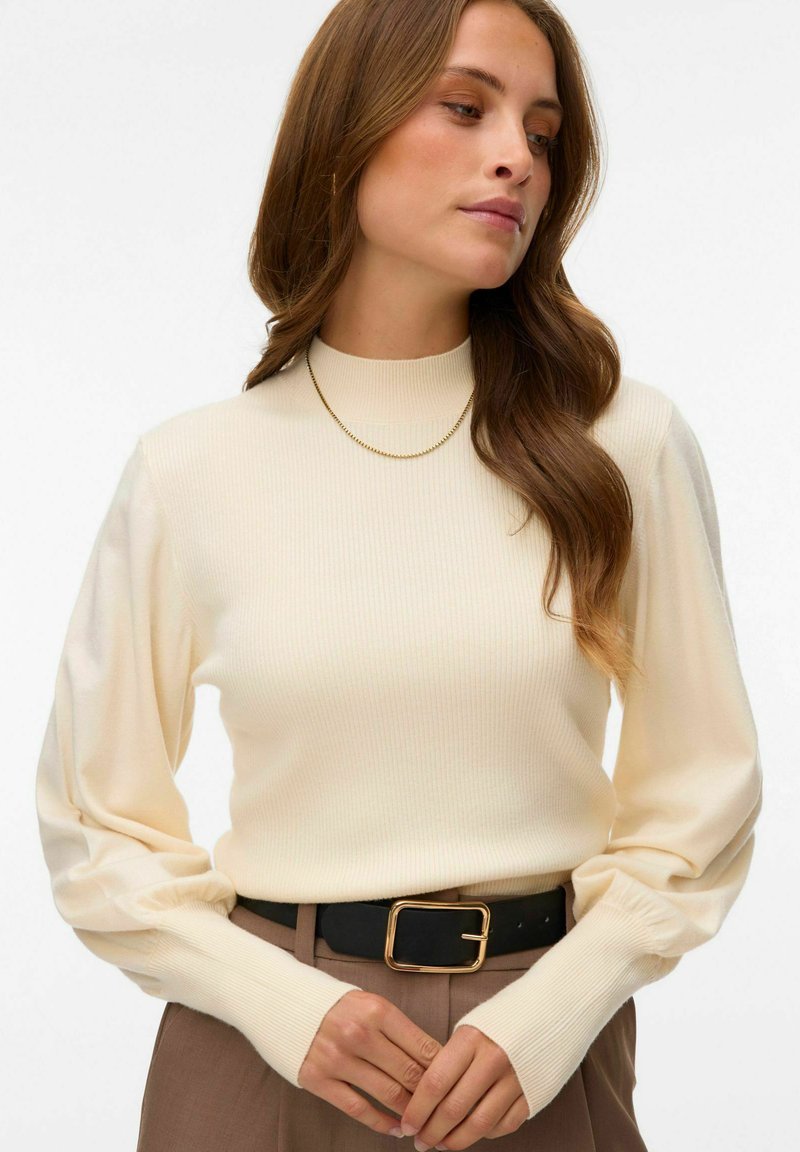 Femme portant un pull côtelé crème à col montant avec des manches bouffantes, un pantalon marron, une ceinture noire avec une boucle dorée, et un collier fin en or.
