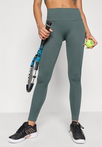Leggings verdes canelados com uma cintura alta, acompanhados de uma raquete de ténis e uma bola. Sapatos desportivos pretos com detalhes em rosa dourado visíveis.