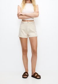 Shorts en denim beige avec ourlets retroussés, dotés d'une fermeture à bouton et à zip à l'avant. Associés à un crop top blanc texturé et des sandales à imprimé léopard.