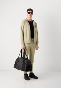 Felpa verde oliva chiaro e pantaloni jogger, t-shirt nera, sneakers nere e occhiali da sole; tiene in mano una borsa da viaggio nera con un logo.