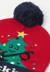 Czerwona dziana czapka z niebieskim pomponem. Ozdobiona zielonym wzorem choinki z gwiazdą, kolorowymi wzorami i dekoracyjnymi cekinami.