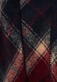 Couverture en laine avec un motif à carreaux dans des tons marine, rouge et beige. Les bords sont dotés de coutures sombres ; texture douce et pelucheuse.