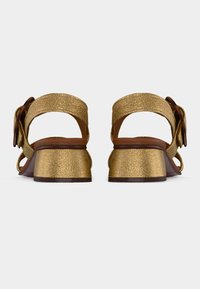 Χρυσά υφασμάτινα πέδιλα με χαμηλό τακούνι, λουριά στο αστράγαλο και μεγάλες στρογγυλές αγκράφες, φωτογραφημένα από το πίσω μέρος σε απλό φόντο.