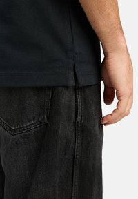 Camiseta de algodón negra con una abertura lateral, combinada con pantalones de mezclilla gris oscuro que presentan bolsillos visibles y un acabado texturizado.