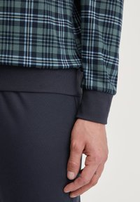 Langarm gestreepte blouse in donkergroen en marineblauw met witte accenten, gecombineerd met een aansluitende donkergrijze manchetten en tailleband, die een gestructureerde stof laat zien.