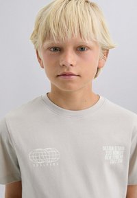 T-shirt en coton gris clair avec un graphique de globe et du texte sur la poitrine gauche. Encolure ras du cou, manches courtes, texture lisse.