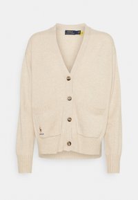 Béžový cardigan s výstřihem do V, žebrovanými manžetami a dvěma předními kapsami. Má pět hnědých knoflíků a malé vyšívané logo.