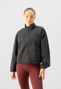 Schwarze Zip-Up-Jacke mit Stehkragen, ausgestattet mit einer Fronttasche und elastischen Bündchen. Der Stoff hat eine glatte Textur und ein kürzeres Design.