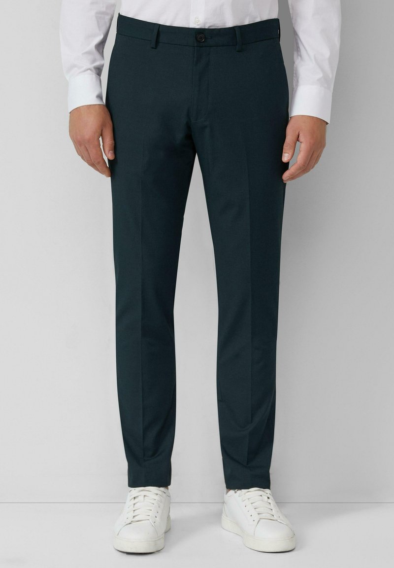 Pantalons taillés vert foncé avec une texture lisse, comprenant une fermeture à bouton à l'avant et des poches latérales, associés à des baskets blanches.
