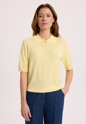 Kobieta ubrana w bladożółtą koszulkę polo z krótkim rękawem i ciemnoniebieskie spodnie, stojąca z jedną ręką w kieszeni na jasnym tle.