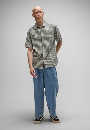 Uomo con camicia a maniche corte a quadri grigia, jeans blu larghi e scarpe da ginnastica nere, in piedi davanti a uno sfondo chiaro e semplice.