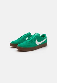 Zaļas sporta kurpes no zamšāda, ar baltu Nike logo, šņorēšanas dizainu un brūnu gumijas zoli ar teksturētu apdari.
