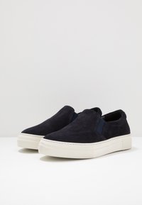 Sapatos slip-on de camurça azul-marinho com detalhes elásticos nas laterais, sola de borracha branca e costuras, apresentando uma textura suave e perfil baixo.