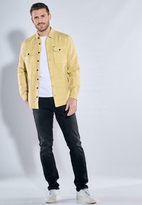 Gelbes Hemd mit Knöpfen und zwei Brusttaschen, kombiniert mit schwarzen Jeans und weißen Turnschuhen. Glattes Material, strukturierte Passform und minimalistisches Design.