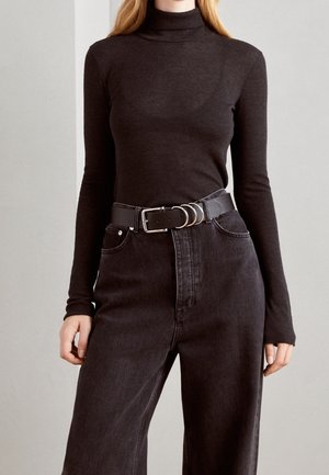 Kobieta ubrana w dopasowany ciemnobrązowy golf włożony do wysokich czarnych dżinsów z czarnym paskiem z srebrnymi kółkami.