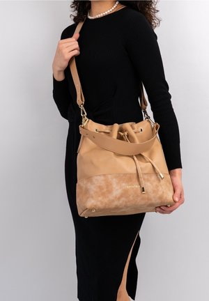 Ruskeanahkainen säilytyskori, jossa on mokkapohja, nyörisuljenta ja kultasävyiset metalliosat. Säädettävä olkahihna ja logoyksityiskohta edessä.