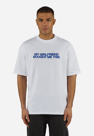 Nuori mies, jolla on lyhyet hiukset ja valkoinen t-paita, jossa lukee sinisellä tekstillä "MY GIRLFRIEND BOUGHT ME THIS", seisoo yksinkertaisen taustan edessä.