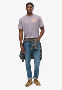 Camiseta de algodón de color púrpura claro con gráfico naranja, jeans azules remangados en los puños y una camisa de franela a cuadros azul marina atada a la cintura. Zapatos marrones.