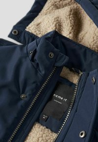 Chaqueta exterior de color azul marino con un suave forro de felpa beige, que cuenta con una cremallera frontal, botones a presión y una etiqueta para la identificación de la marca.