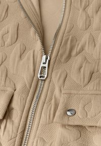 Beige vadderad jacka med en silverdragkedja, strukturerade mönster och en bröstficka med en metallknapp.