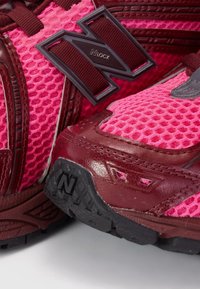 Chaussures de sport roses et bordeaux avec tissu en mesh, semelles en caoutchouc et grands accents du logo "N" sur les côtés et la zone des orteils.