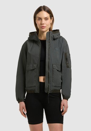 Žena v tmavě šedé bundě s kapucí a několika kapsami přes černý krátký top a černé cyklistické šortky na jednobarevném pozadí.