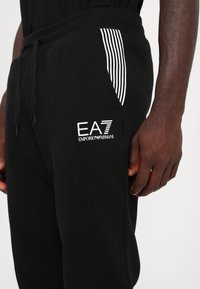 Svarta sweatpants i ett texturerat tyg, med en vit logotyp och en randig fickdetalj. Elastisk midja och justerbar dragsko.
