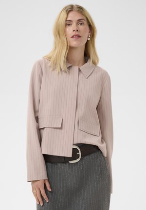 Mujer con cabello rubio hasta los hombros que lleva una chaqueta rosa claro de rayas finas y una falda gris de rayas finas con un cinturón negro.