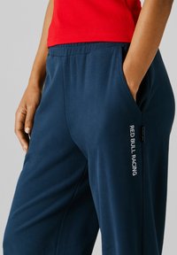 Osoba ubrana w granatowe spodnie dresowe z napisem "RED BULL RACING" na udzie oraz czerwoną bluzę, z ręką włożoną do lewej kieszeni.